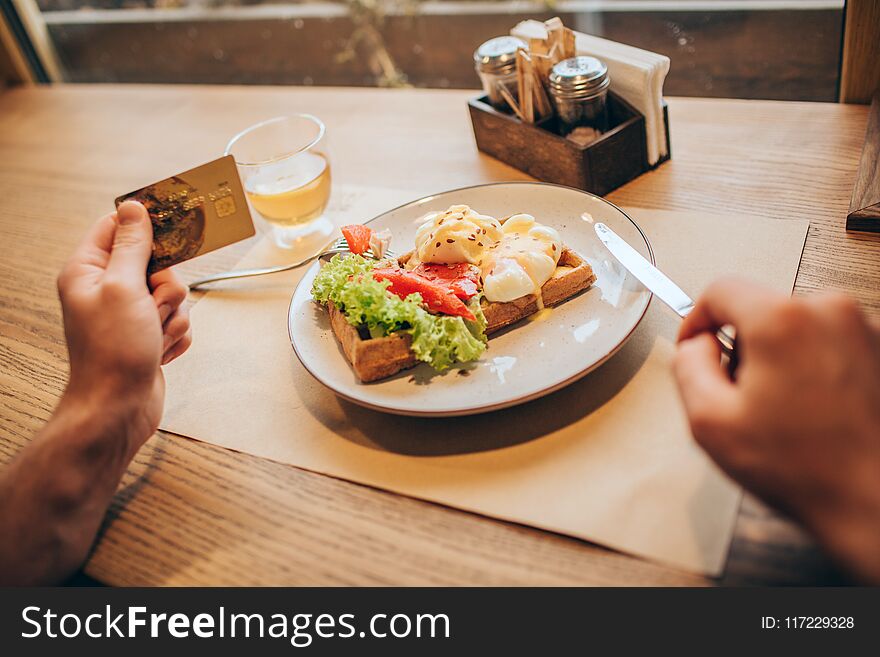 A picture of guy`s hands holding gold card in left one. He is holding a knife with right one. There is a plate with tasty food on table. Close up