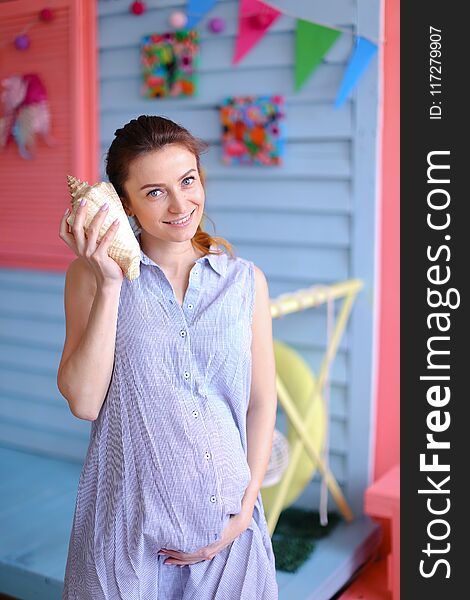 Young nice pregnant woman holding seashell to ear and standing near children house. Concept of pregnance and oceanic theme.