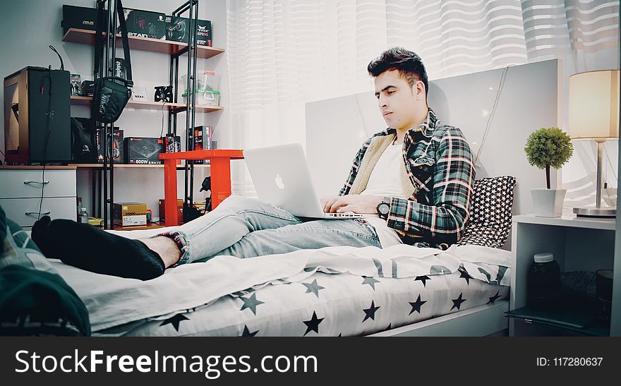 Man in Green, Blue, and Black Plaid Sports Shirt Sitting on Bed Using Silver Macbook