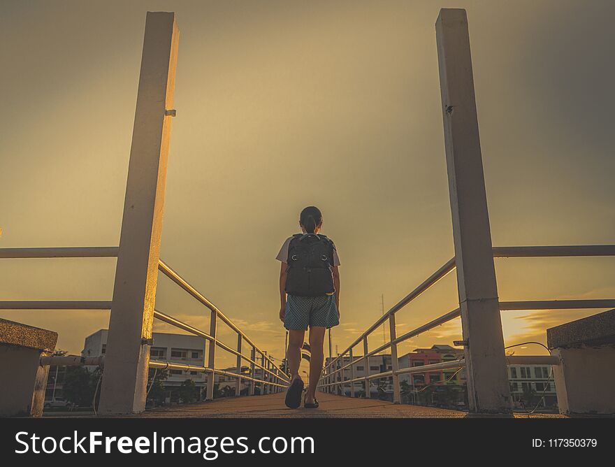 Asian Woman tourist walking alone with backpack on the bridge in the city in the evening on sunset with yellow sky. Walk away
