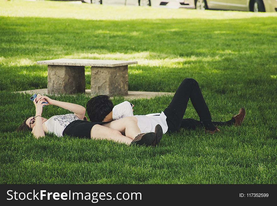 Man And Woman Laying On Green Grass Near Concrete Bench