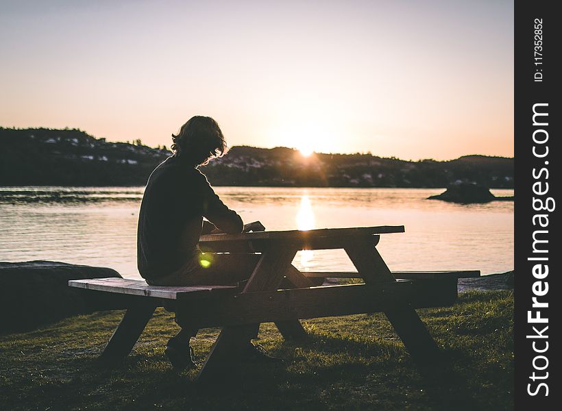 Silhouette Of Person In Black Top Sitting On Picnic Bench Near Body Of Water During Sunset