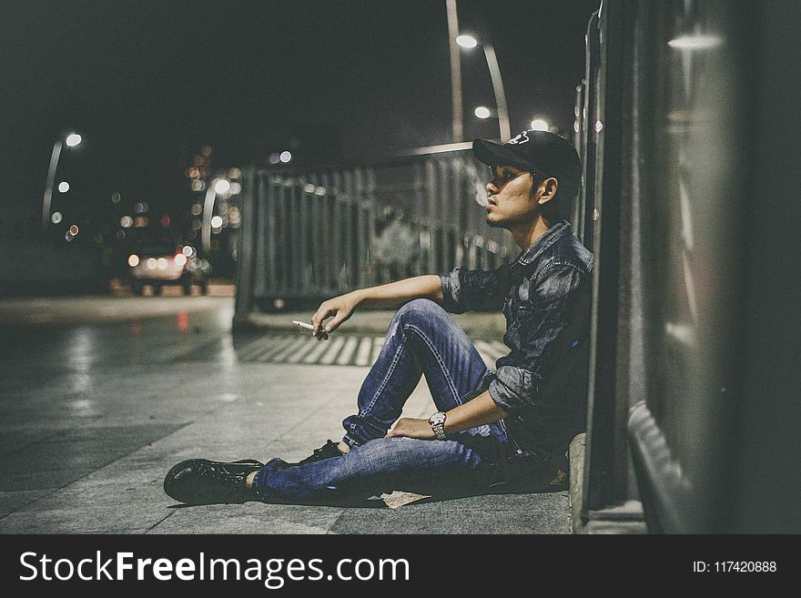 Shallow Focus Photography of Man Sitting Beside Railing Holding Cigarette Stick