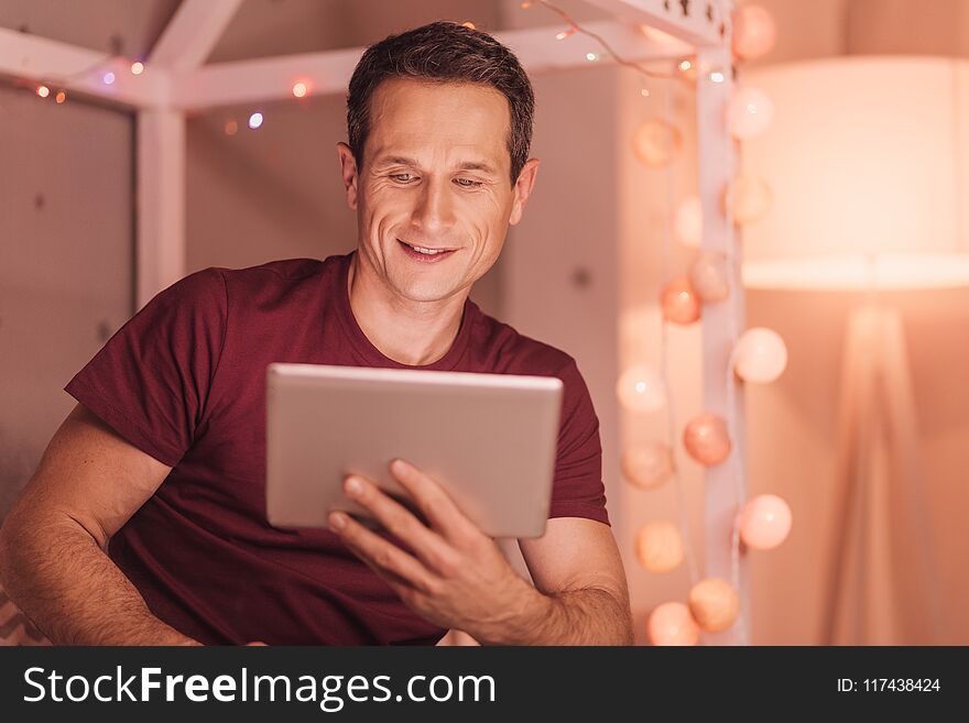 Modern entertainment. Cheerful positive young man smiling and looking at the tablet screen while relaxing at home. Modern entertainment. Cheerful positive young man smiling and looking at the tablet screen while relaxing at home