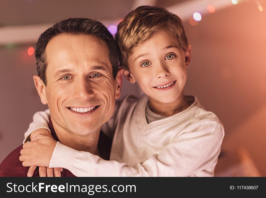 Men in the family. Nice pleasant cute boy smiling and hugging his father while looking at you. Men in the family. Nice pleasant cute boy smiling and hugging his father while looking at you