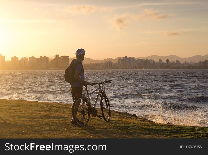 Man With Bicycle Standing On Cliff Front Of Water Waves Golden Hour Photography