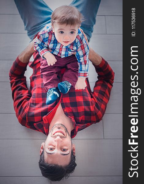 Lying Man Wearing Red and Black Gingham Long-sleeved Shirt Holding Toddler Sitting on His Stomach