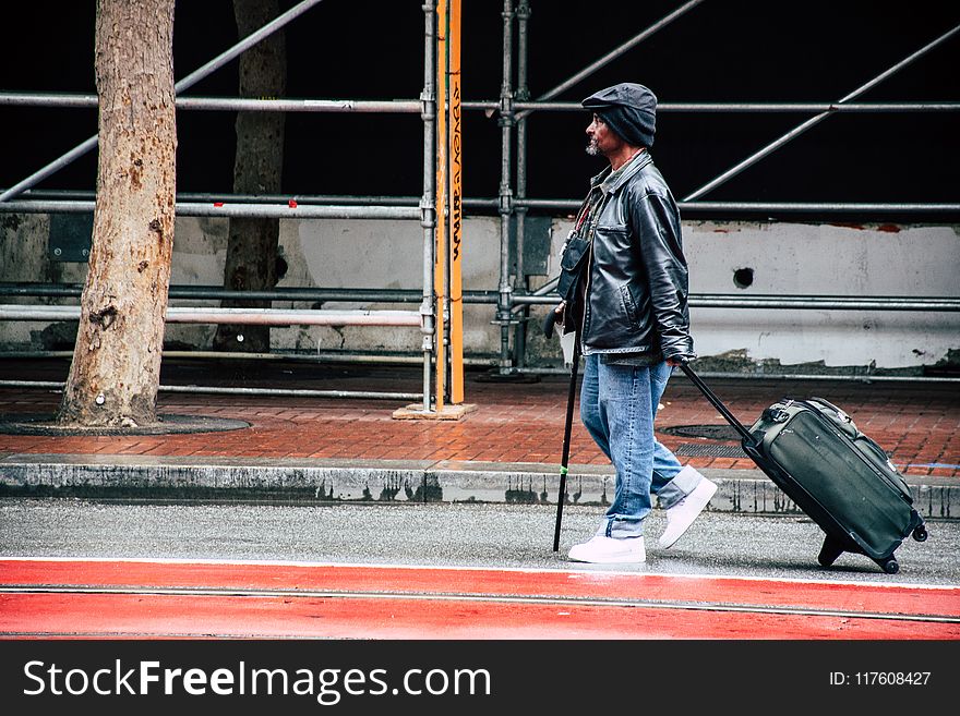 Walking Man in Black Leather Jacket and Blue Denim Pants Holding Luggage Bag
