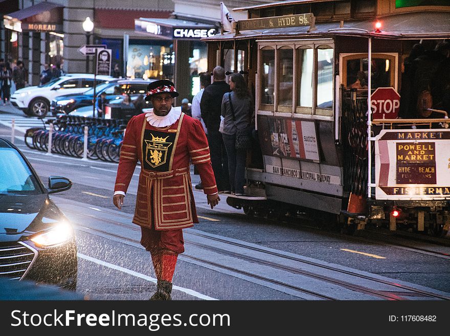 Man Wearing Red and Yellow Dress Walking on Concrete Road Near White and Red Cable Train