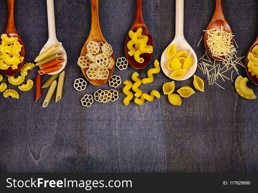 Different pasta in spoons on a dark wooden background, top view.