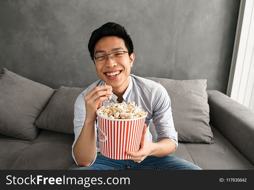 Portrait of a cheerful young asian man eating popcorn while sitting on a couch at home and watching TV