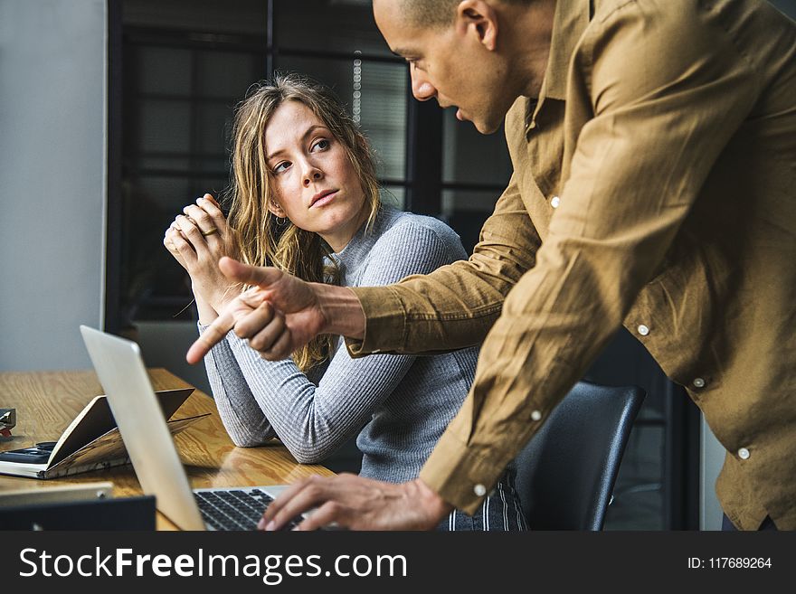 Man in Brown Long-sleeved Button-up Shirt Standing While Using Gray Laptop Computer on Brown Wooden Table Beside Woman in Gray Long-sleeved Shirt Sitting