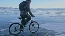 Man Is Riding A Bicycle On Ice. The Cyclist Is Dressed In A Gray Down Jacket, Backpack And Helmet. Ice Of The Frozen Stock Photo Man Is Riding A Bicycle On Ice. The Cyclist Is Dressed In A Gray Down Jacket, Backpack And Helmet. Ice Of The Frozen Stock Photo