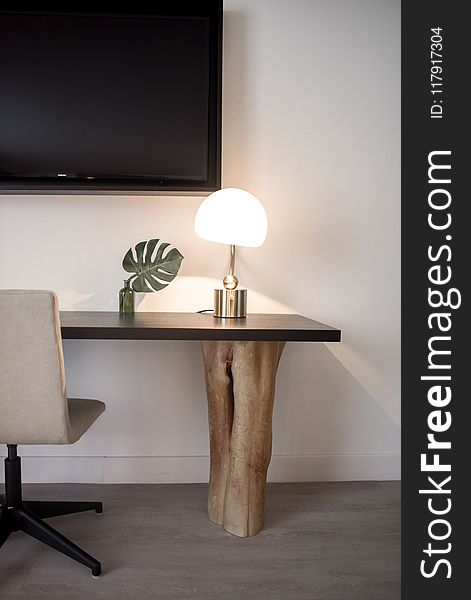 Stainless Steel Base White Shade Table Lamp on Brown Wooden Desk Near White Painted Wall With Wall Mounted Flat Screen T V