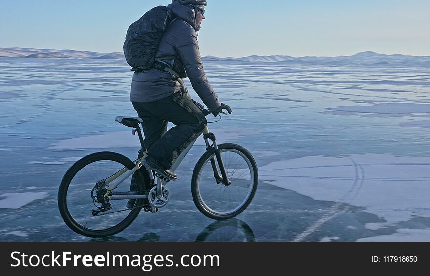 Man Is Riding A Bicycle On Ice. The Cyclist Is Dressed In A Gray Down Jacket, Backpack And Helmet. Ice Of The Frozen