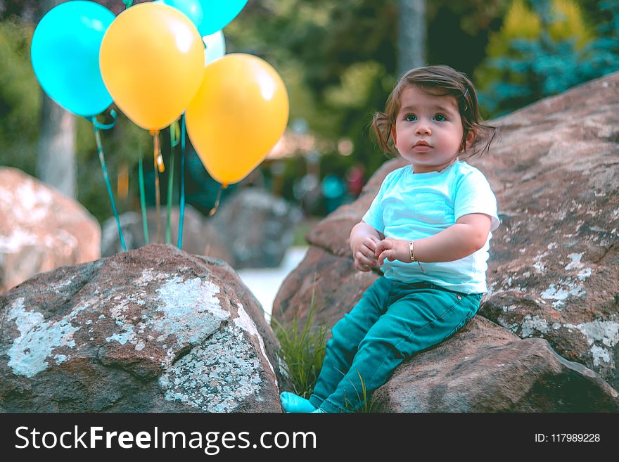 Toddler Wearing White Shirt Sitting on Rock Beside Yellow and Blue Balloons