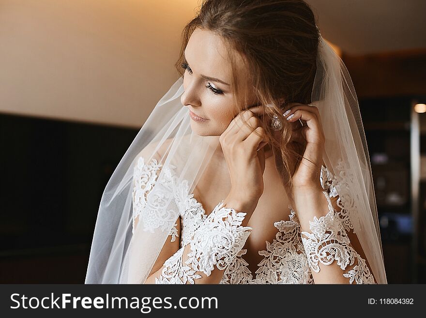 Portrait of beautiful and stylish model girl, young bride with professional makeup in lace dress puts on earring and posing in interior.