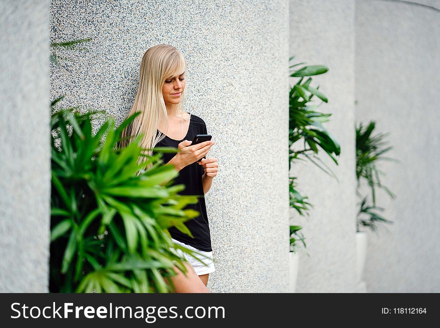 Woman Wearing Black Scoop-neck Shirt Standing in Front on Concrete Column Holding Smartphone