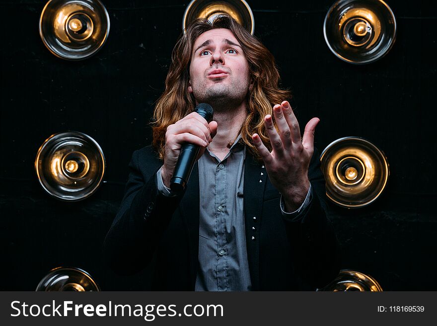 Brutal singer with microphone on the stage, with long hair, against the background of bright lanterns, scene. Brutal singer with microphone on the stage, with long hair, against the background of bright lanterns, scene