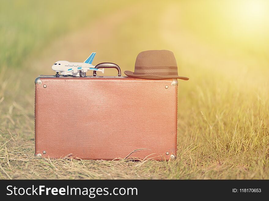 Travel and adventure concept. Vintage brown suitcase and hat with toy airplane near the road in the green field.