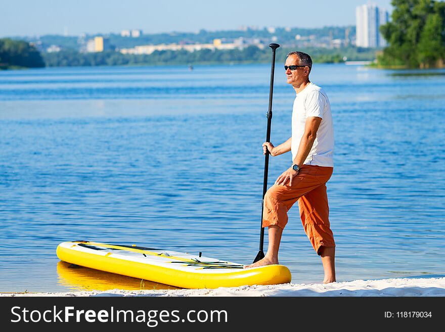 Adult man in sunglasses is standing next to SUP board on the beach on the cityscape background. Stand up paddle boarding - awesome active recreation during vacation.