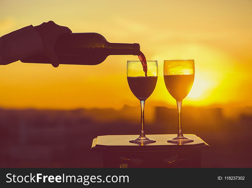 Female hand with bottle pours red wine into glasses on a sunset background.
