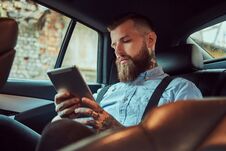 Old-fashioned Tattooed Hipster Guy In A Shirt With Suspenders, Using A Tablet While Sitting In A Luxury Car On Back Seat Stock Image