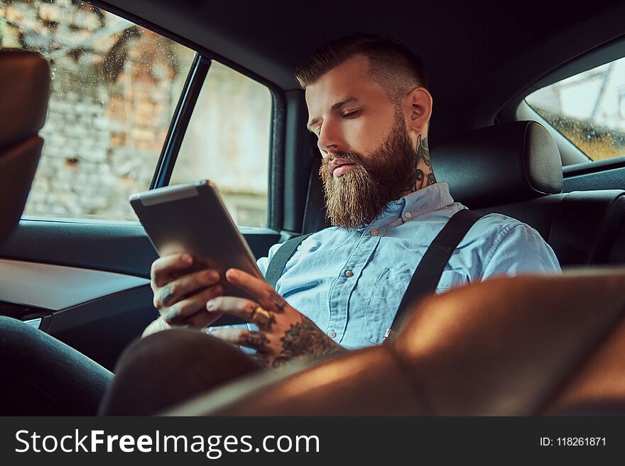 Old-fashioned tattooed hipster guy in a shirt with suspenders, using a tablet while sitting in a luxury car on back seat