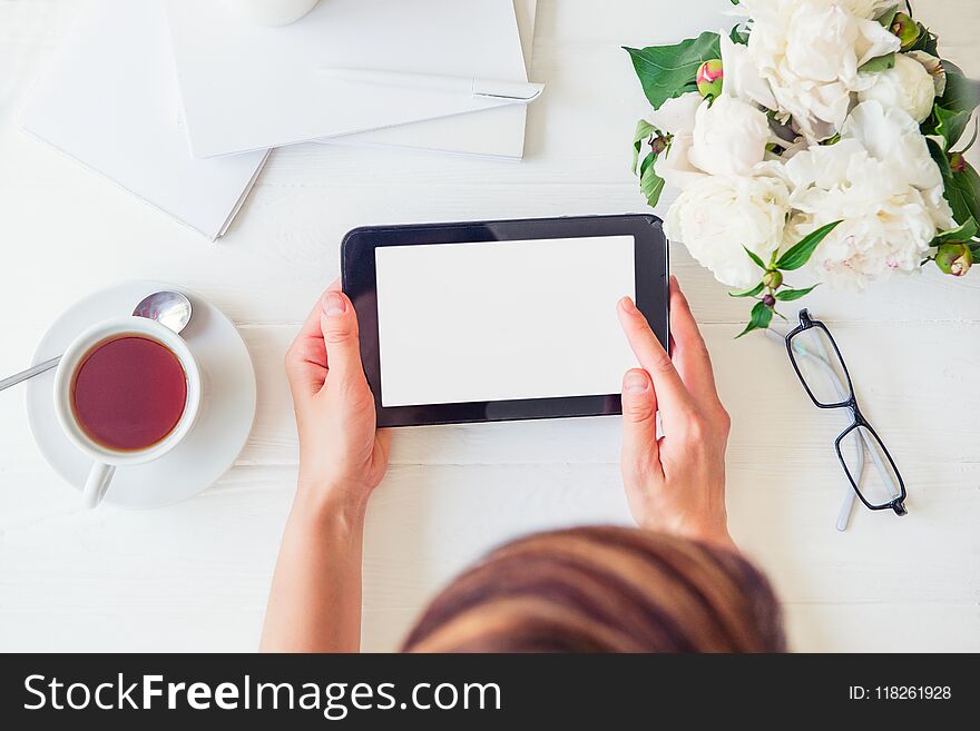 Workspace with girl`s hands holding tablet with blank screen, cup of tea, glasses, notebooks, white peony on woodden table. Top view feminine office desk. Planning day. Freelancer working place