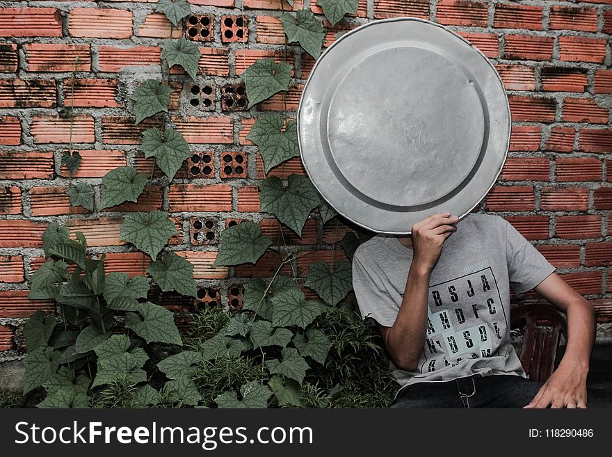 Person Wearing Gray Crew-neck T-shirt holding a tray on the face next to a brick wall