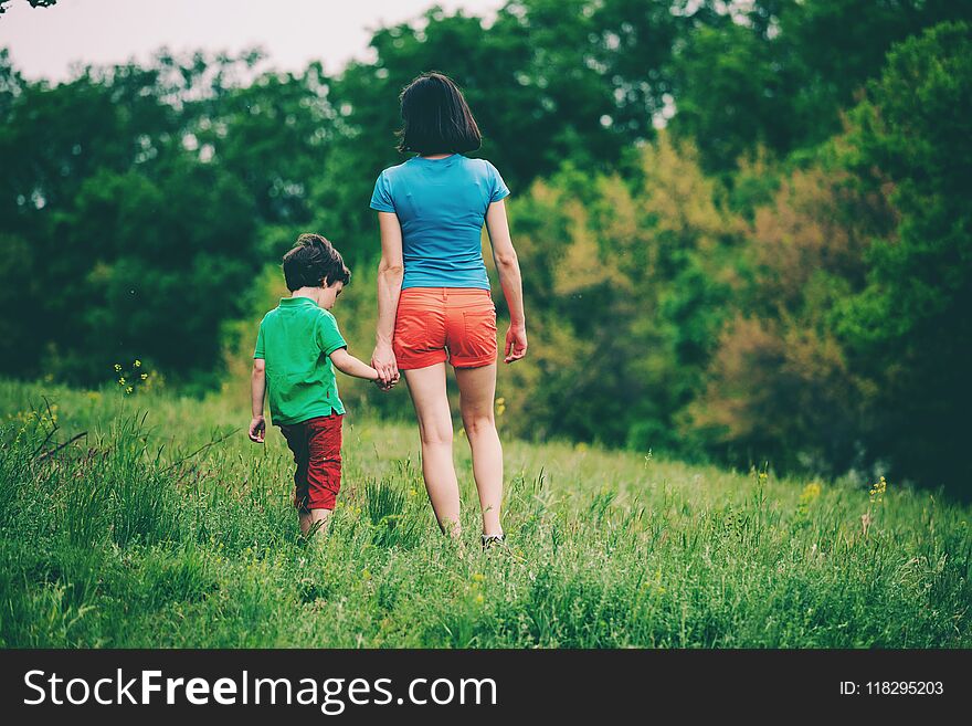The boy walks with his mother in the meadow. A women holds her son by the hand. The kid and mom are walking along the field. Green grass.