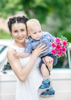 Portrait Of Pretty Young Female Bride Walking, Holding Baby Boy With Wedding Roses Bouquet And Looking Into Camera At Sunny Summer Royalty Free Stock Photography