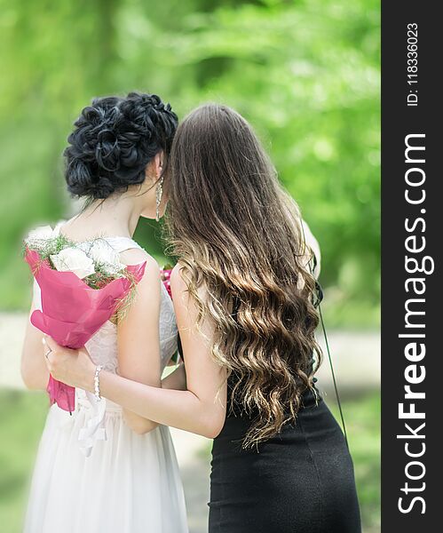 Portrait of back of two young beautiful women hugging each other in green summer park. Pretty females Bride and bridesmaid talking