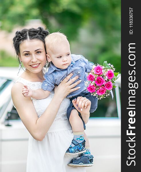Portrait of pretty young female bride walking, holding baby boy with wedding roses bouquet and looking into camera at sunny summer