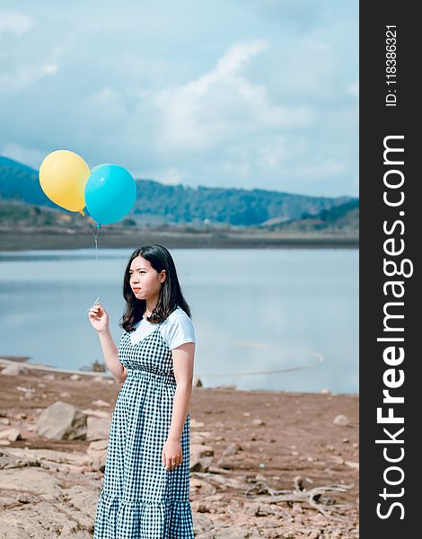 Woman Wearing Blue and White Plaid Dress Holding Blue and Yellow Balloons Near Calm Body of Water at Daytime