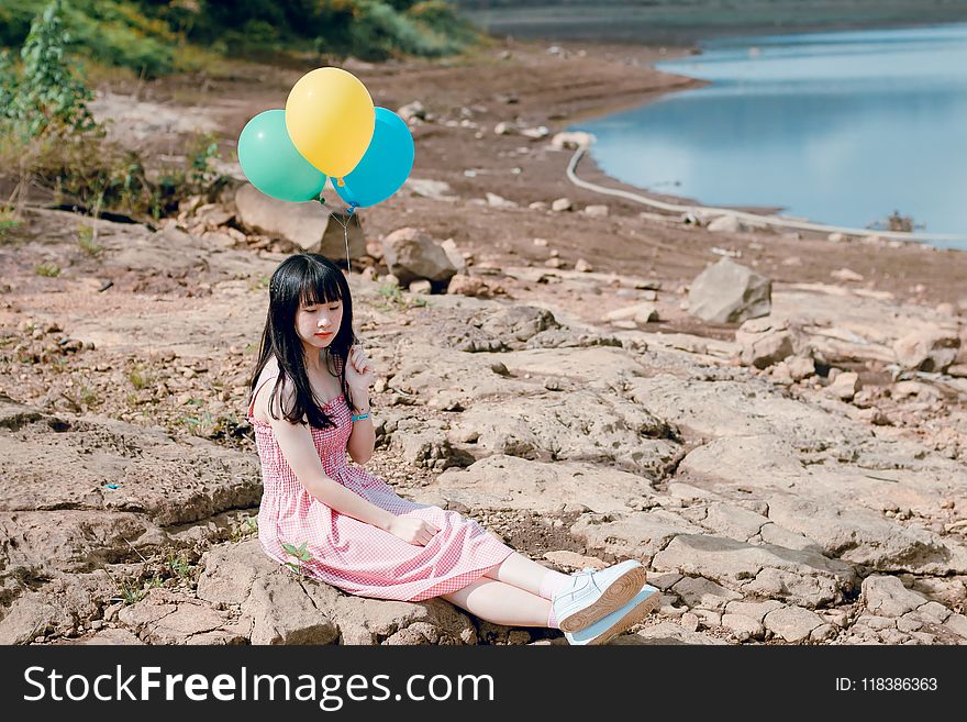 Woman in Pink Thick Strap Dress Holding a Yellow and Blue Balloons While Sitting on Rock Terrain