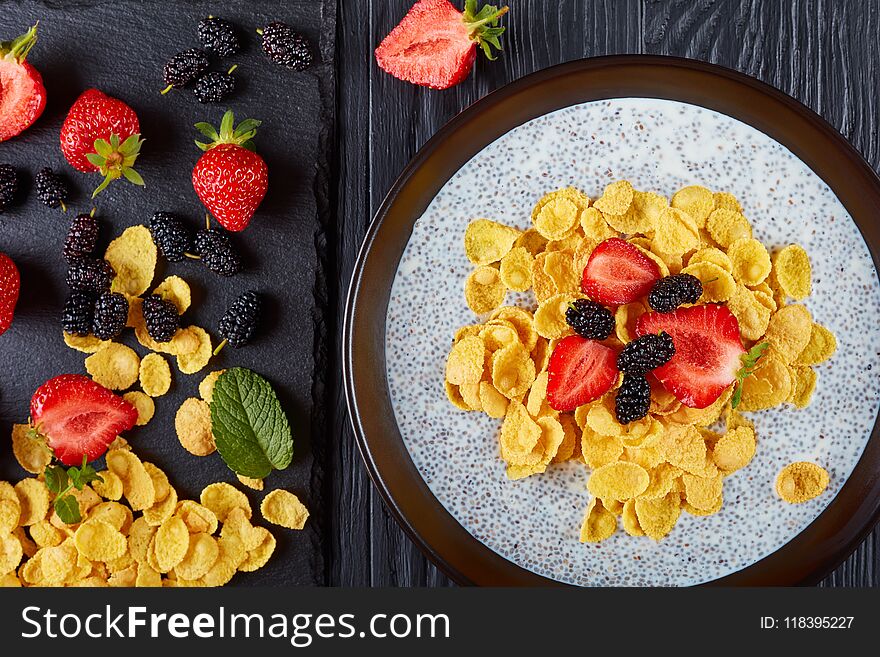 Healthy breakfast - Corn flakes with chia seeds pudding, fresh strawberry,and mulberries in a bowl on black wooden table with ingredients on cutting board, horizontal view from above, close-up, flat lay