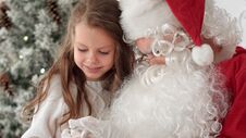 Santa Claus Tying A Ribbon Bow On A Gift For A Little Girl Sitting Near The Christmas Tree At Home Royalty Free Stock Image