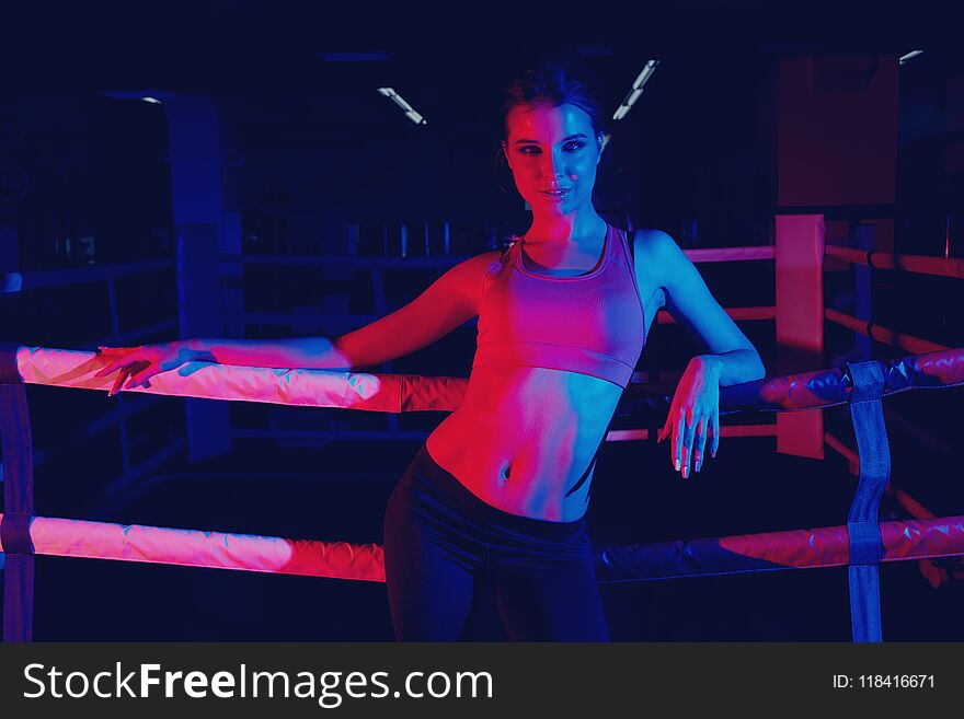 Attractive brunette woman with pony tail, dressed in sportswear, illuminated by dim neon light, sitting and leaning her back against ropes of boxing ring. Beautiful female fitness model posing at gym.