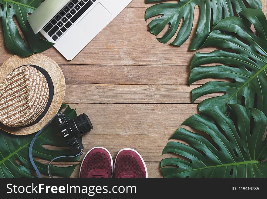 Flat lay creative frame of tropical nature leaves Monstera on rustic wood grunge background with laptop computer, retro camera, hat and pair of sneaker shoes, digital nomad and life balance concepts.