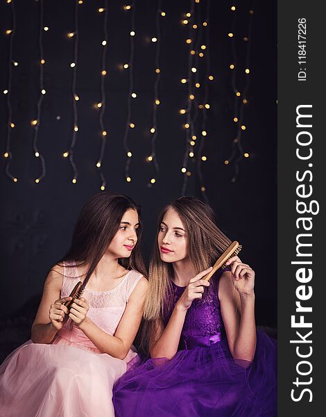 Cheerful girlfriends. Two girls comb each other`s hair, sitting side by side in studio on dark background. Copy space.