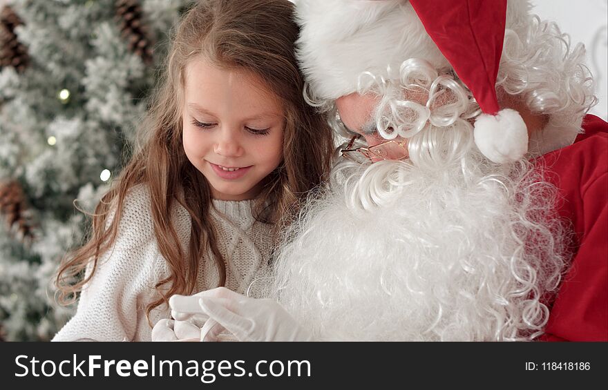 Santa Claus tying a ribbon bow on a gift for a little girl sitting near the Christmas tree at home.