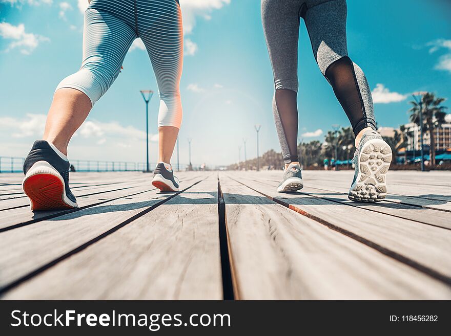 Low angle close up of female feet running along the wooden path. Two girls are training near the sea. Low angle close up of female feet running along the wooden path. Two girls are training near the sea
