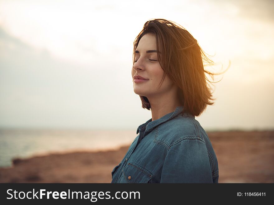 Great pleasure. Charming calm young woman is standing near sea with closed eyes and expressing delight. She is posing against wonderful sunset while enjoying last rays of the sun. Great pleasure. Charming calm young woman is standing near sea with closed eyes and expressing delight. She is posing against wonderful sunset while enjoying last rays of the sun