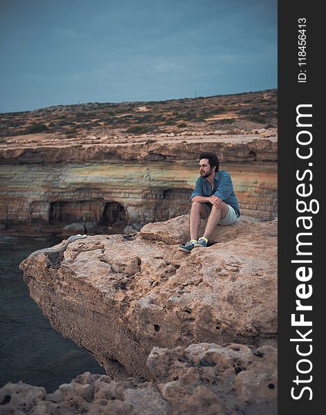 Feeling lonely. Full length of young stylish bearded man is sitting on rocks while looking aside thoughtfully. He is spending time near sea. Copy space in the left side. Feeling lonely. Full length of young stylish bearded man is sitting on rocks while looking aside thoughtfully. He is spending time near sea. Copy space in the left side