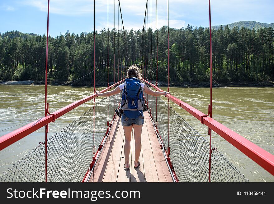 A girl with a backpack walks on a suspension bridge in a clear sunny day. The bridge is stretched over a mountain river. A girl with a backpack walks on a suspension bridge in a clear sunny day. The bridge is stretched over a mountain river.