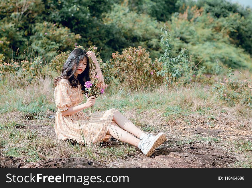 Woman Wearing Brown Dress Sitting on Grasses While Holding Purple Flowers