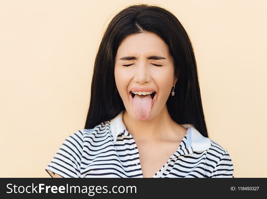 Portrait of beautiful woman makes grimace, keeps mouth wide opened and shows tongue, keeps eyes closed, wears casual clothes, poses against studio background. People and facial expressions concept.