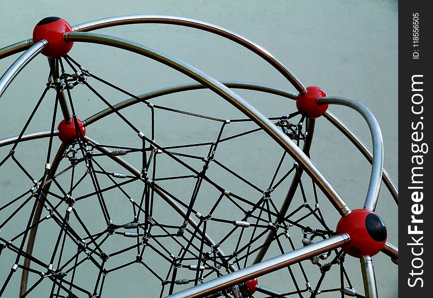 Red plastic ball joints with black vinyl rope mesh on a playground climbing equipment and shiny stainless steel tube framing and aluminum clamps in a public park under blue sky. Red plastic ball joints with black vinyl rope mesh on a playground climbing equipment and shiny stainless steel tube framing and aluminum clamps in a public park under blue sky