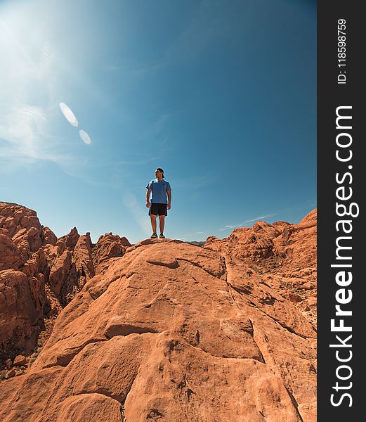 Man Wearing Blue Shirt And Black Shorts Standing On Top Of Brown Rock Formations Under Clear Sky